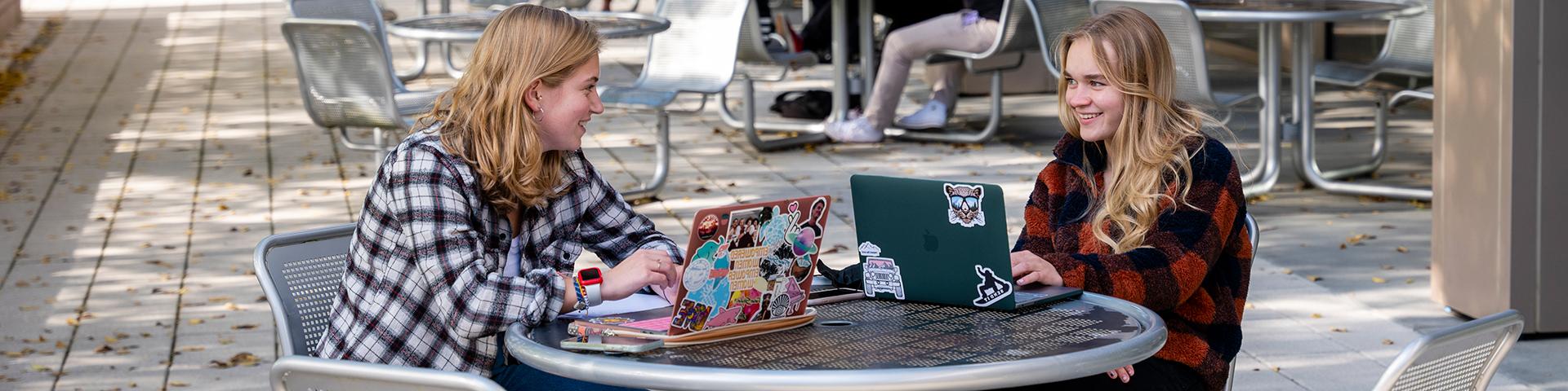 Two female students sitting outside of Gaige on their laptops